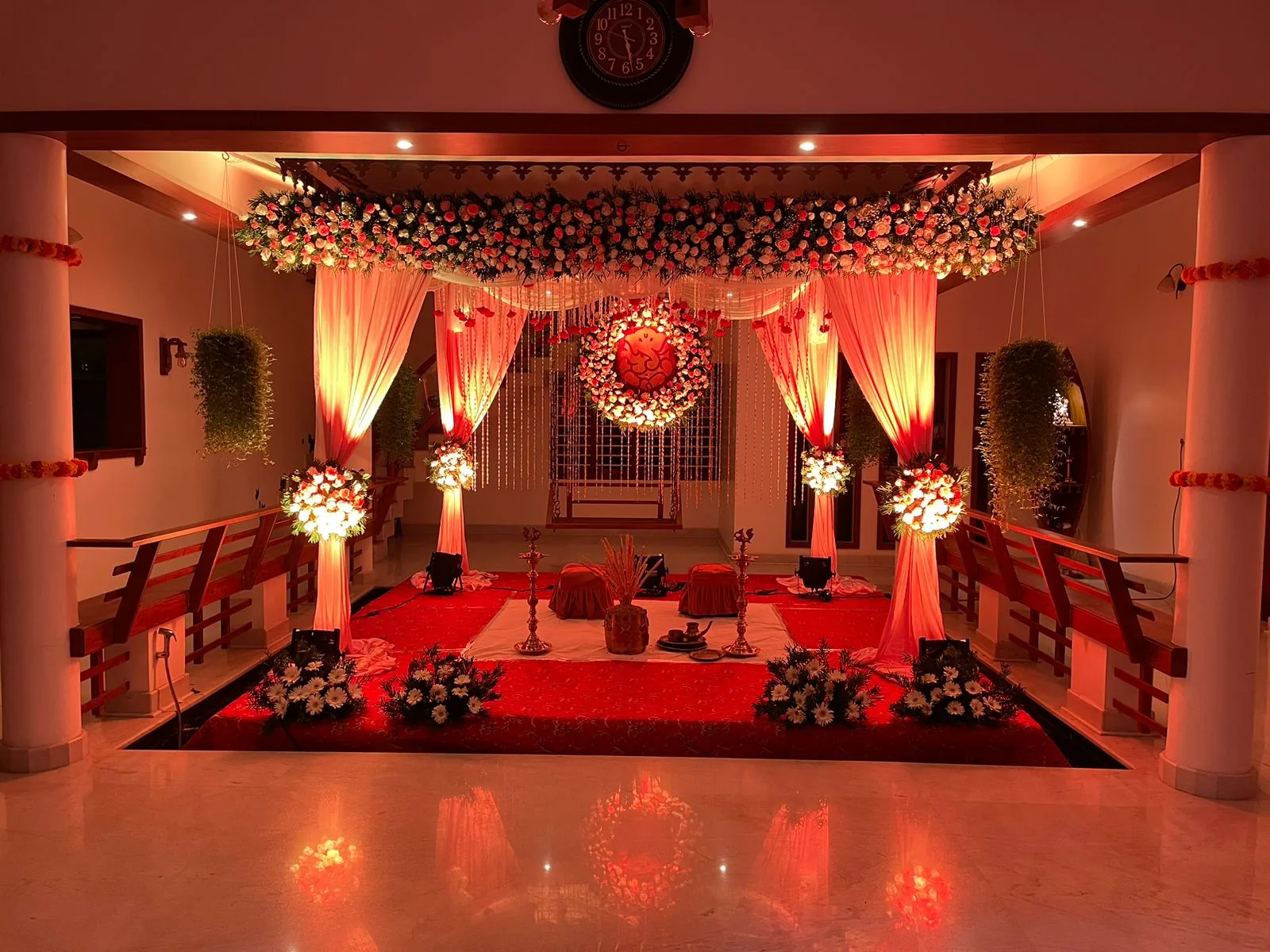 Wedding mandap with draped pillars, roses and brass lamps under warm red light